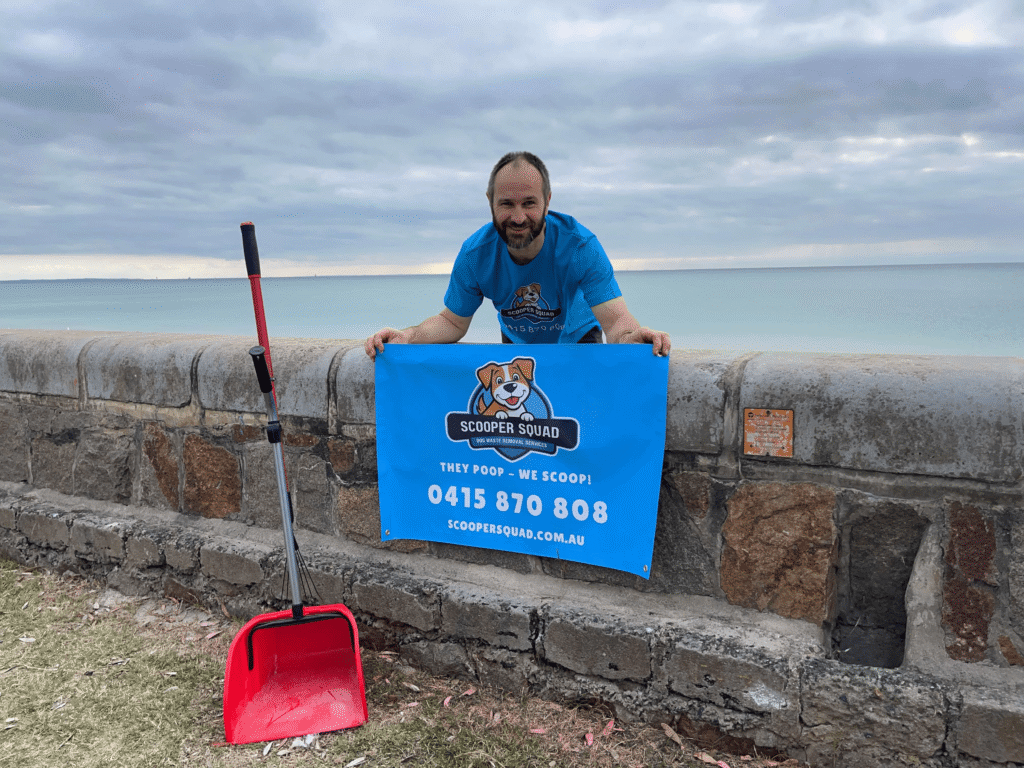 Scooper Squad owner holding blue dog waste removal banner and pooper scooper tools by the beach